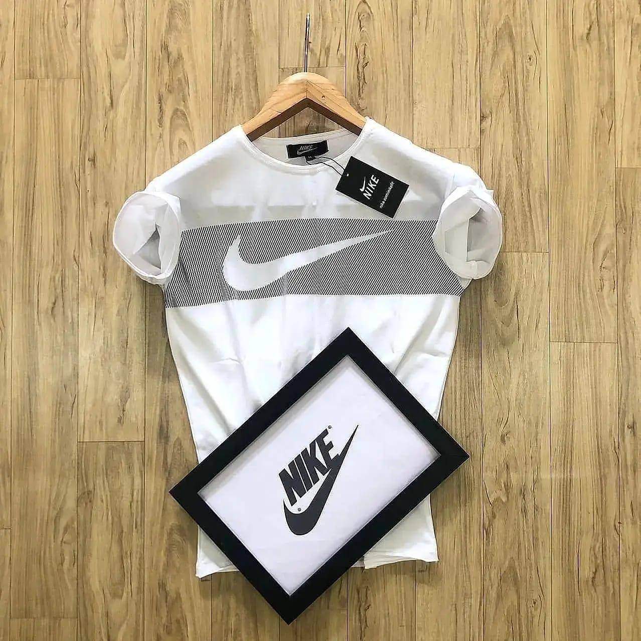 Nike White Men's T-Shirt with a bold swoosh logo across the chest, displayed on a hanger against a wooden background. Includes a Nike-branded tag and framed logo.